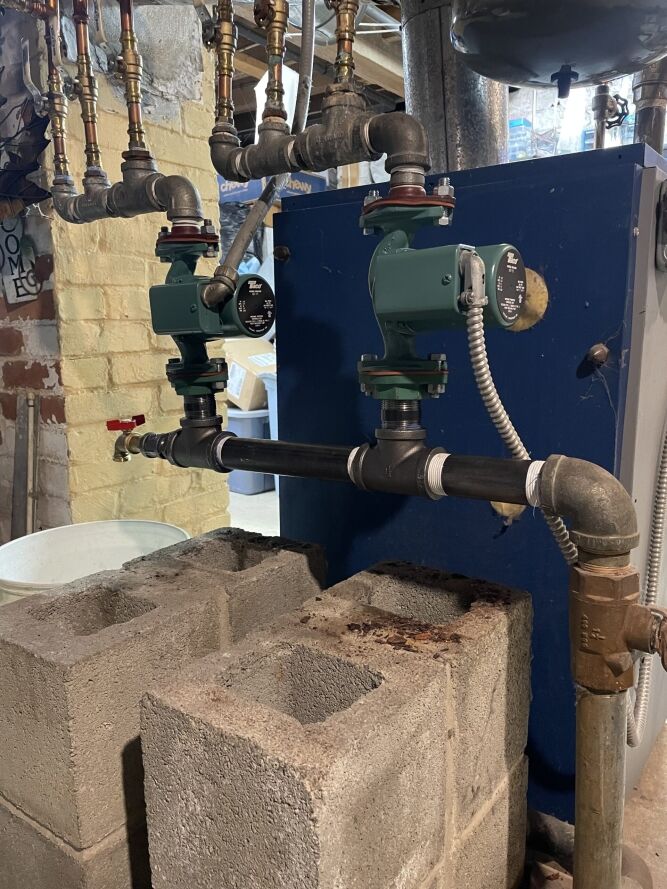 A basement heating system with exposed pipes, two green water pumps, and concrete blocks supporting the piping structure.