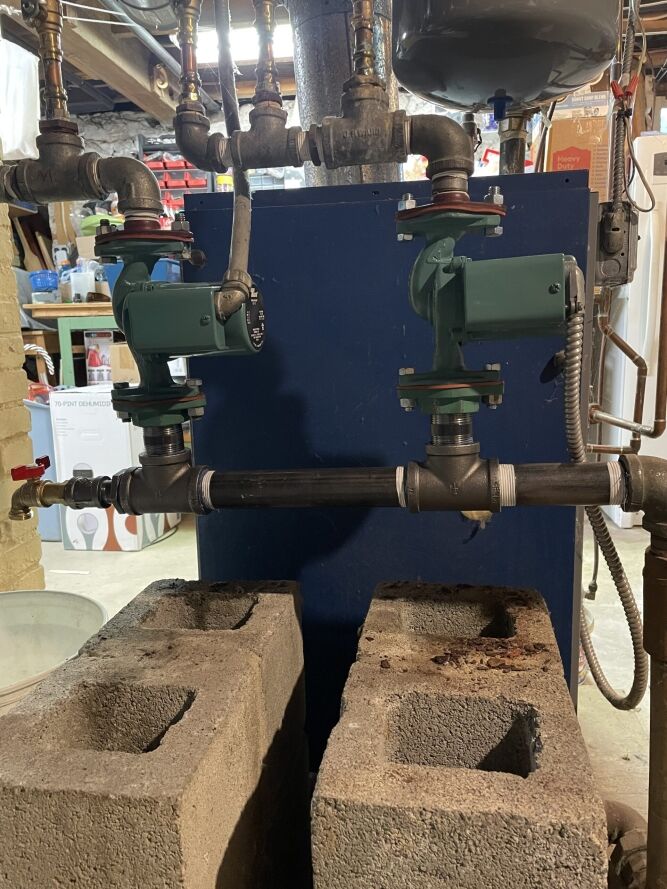 Two green circulation pumps are installed on black pipes, supported by concrete blocks, in a basement utility area with exposed plumbing and various tools in the background.
