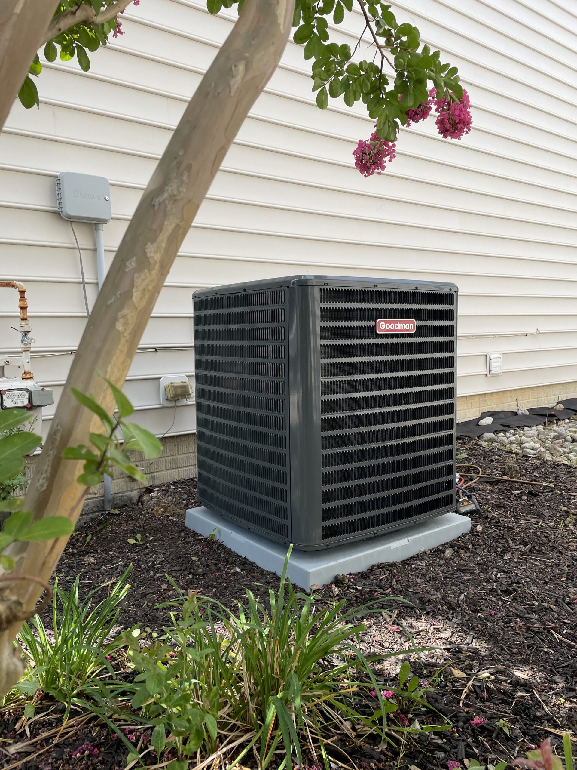 A Goodman air conditioning unit is installed outside a house next to a beige siding wall, with a small tree and plants in the foreground.