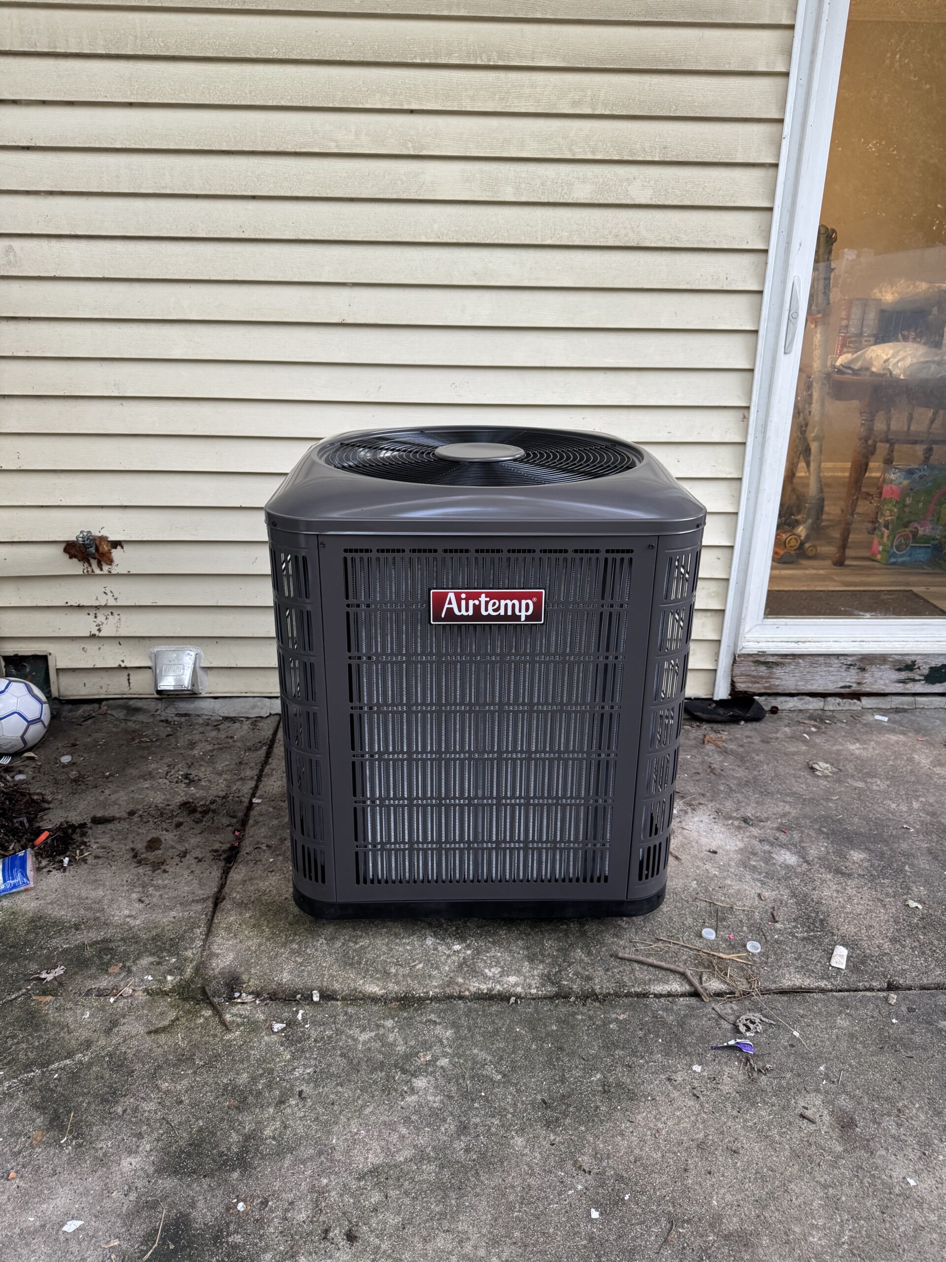 A gray Airtemp outdoor air conditioning unit sits on a concrete patio next to a beige house with vinyl siding and a glass door.