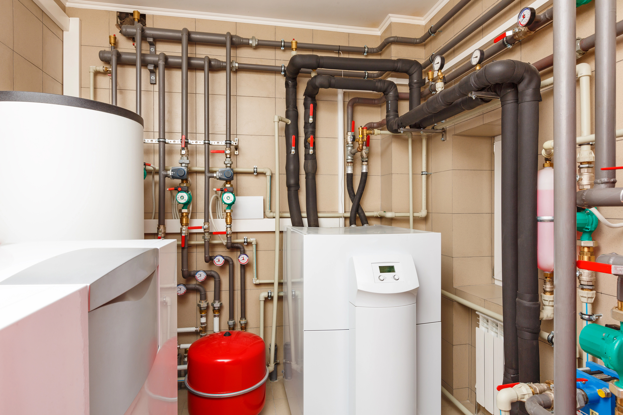A utility room with a modern boiler system, multiple pipes, valves, gauges, a red expansion tank, and beige tiled walls.