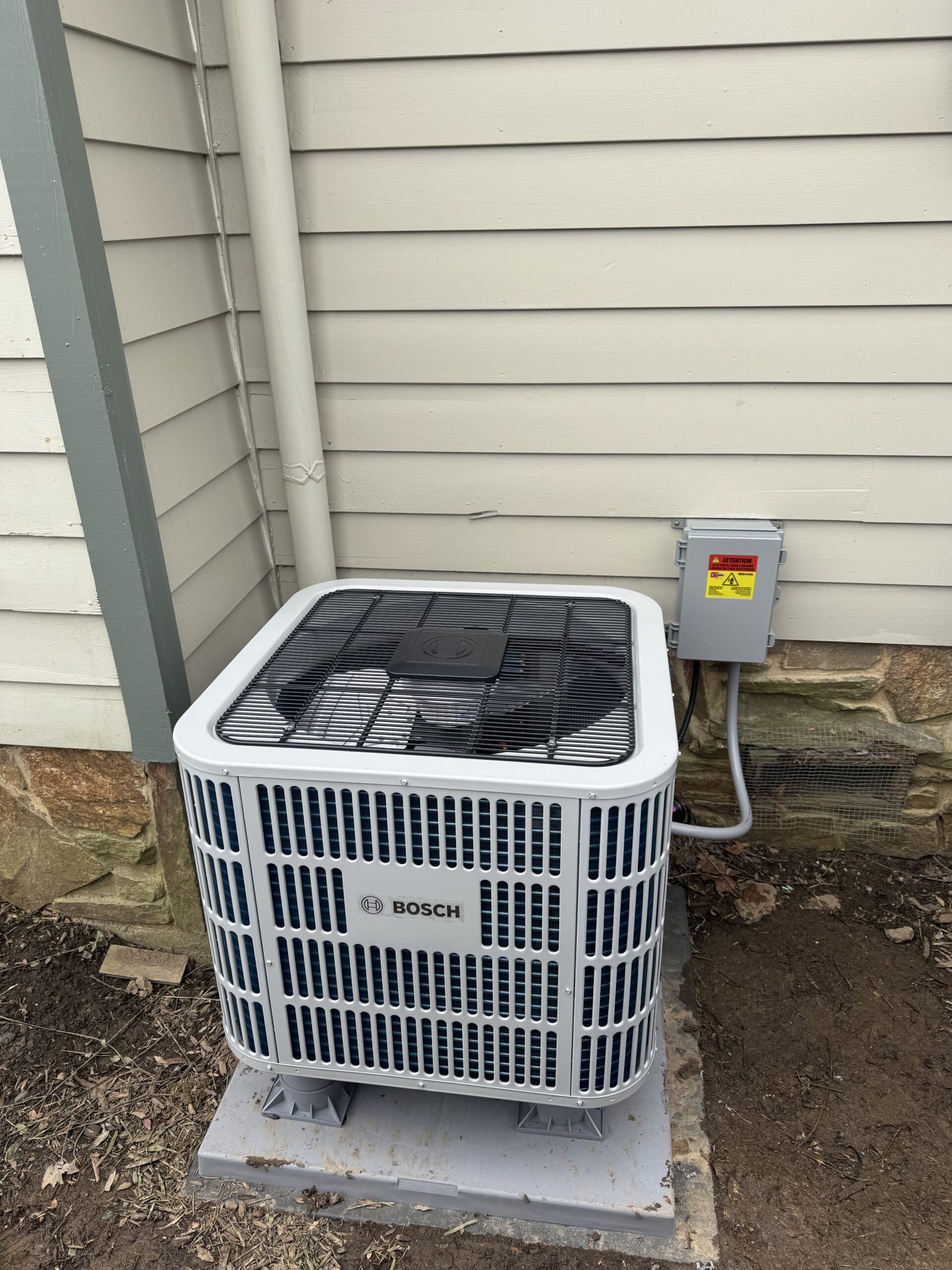 A Bosch outdoor air conditioning unit is installed on a concrete pad next to a house's exterior wall, connected to an electrical box.