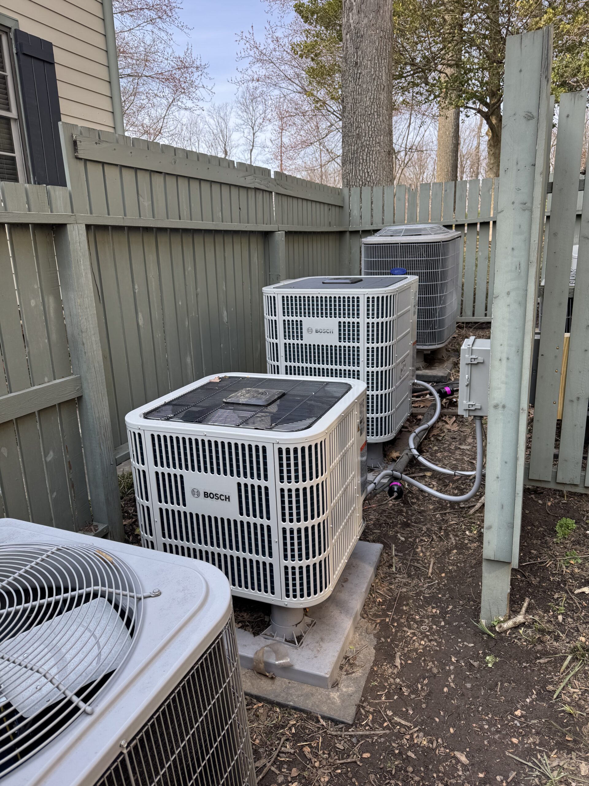 Four outdoor Bosch HVAC units are installed on concrete pads beside a wooden fence and house, connected with pipes and electrical wiring.