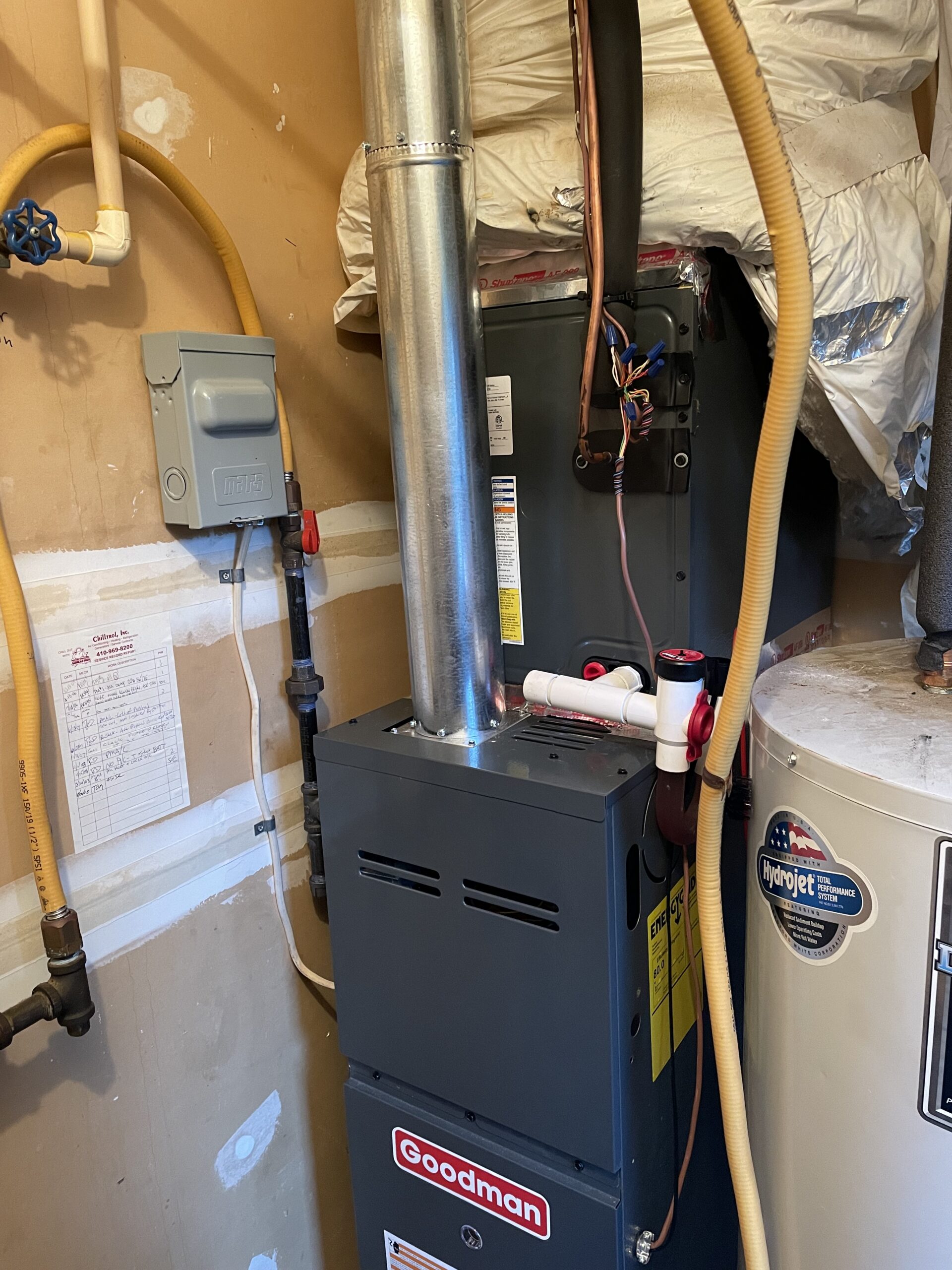 A Goodman furnace installed next to a water heater and various pipes in a utility room with unfinished walls.
