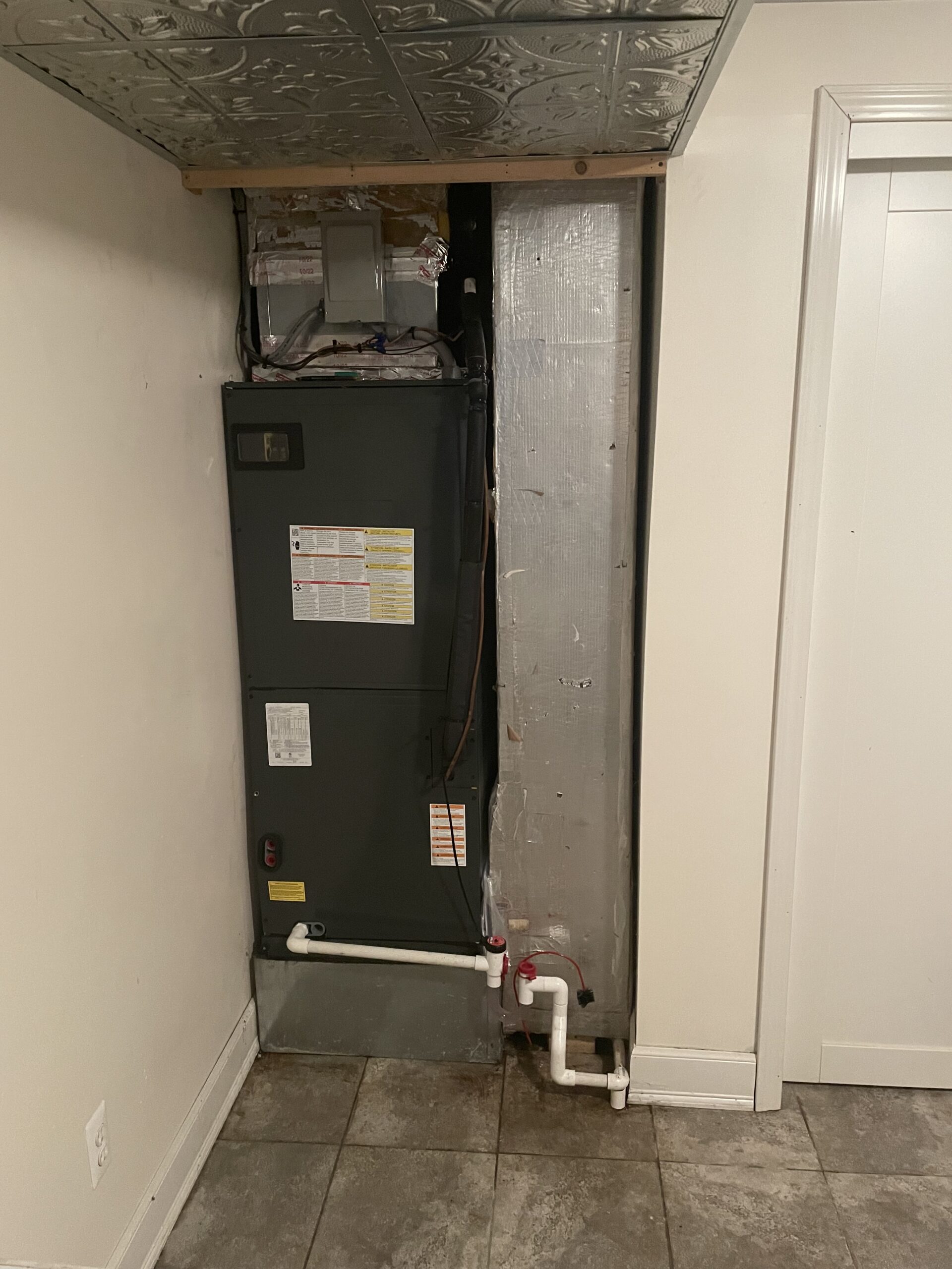An HVAC unit installed in a recessed wall area with visible ducts and pipes, next to a white door and tiled floor.