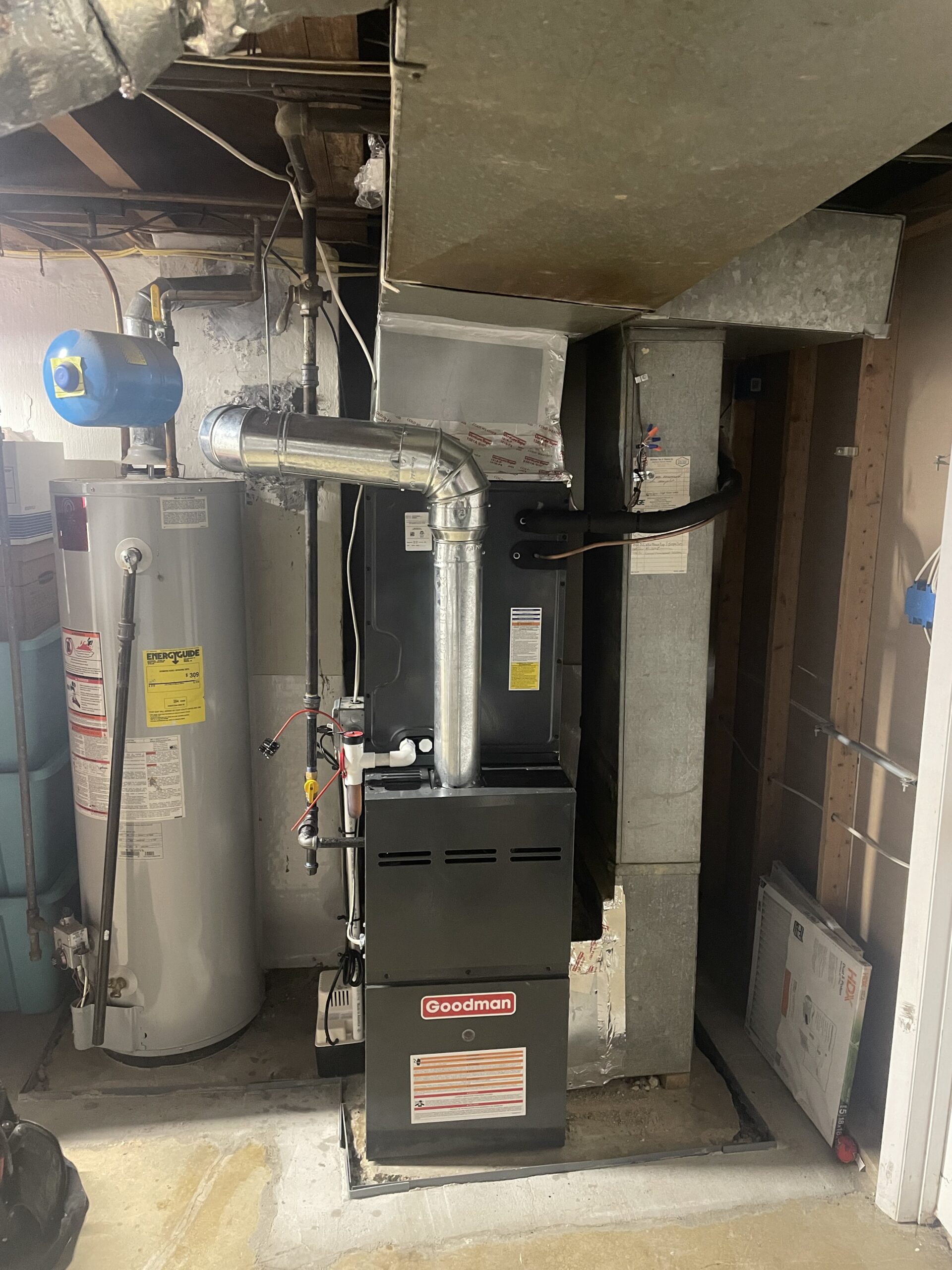 A Goodman furnace unit is installed next to a water heater in a basement utility area, with ductwork and pipes connected above.