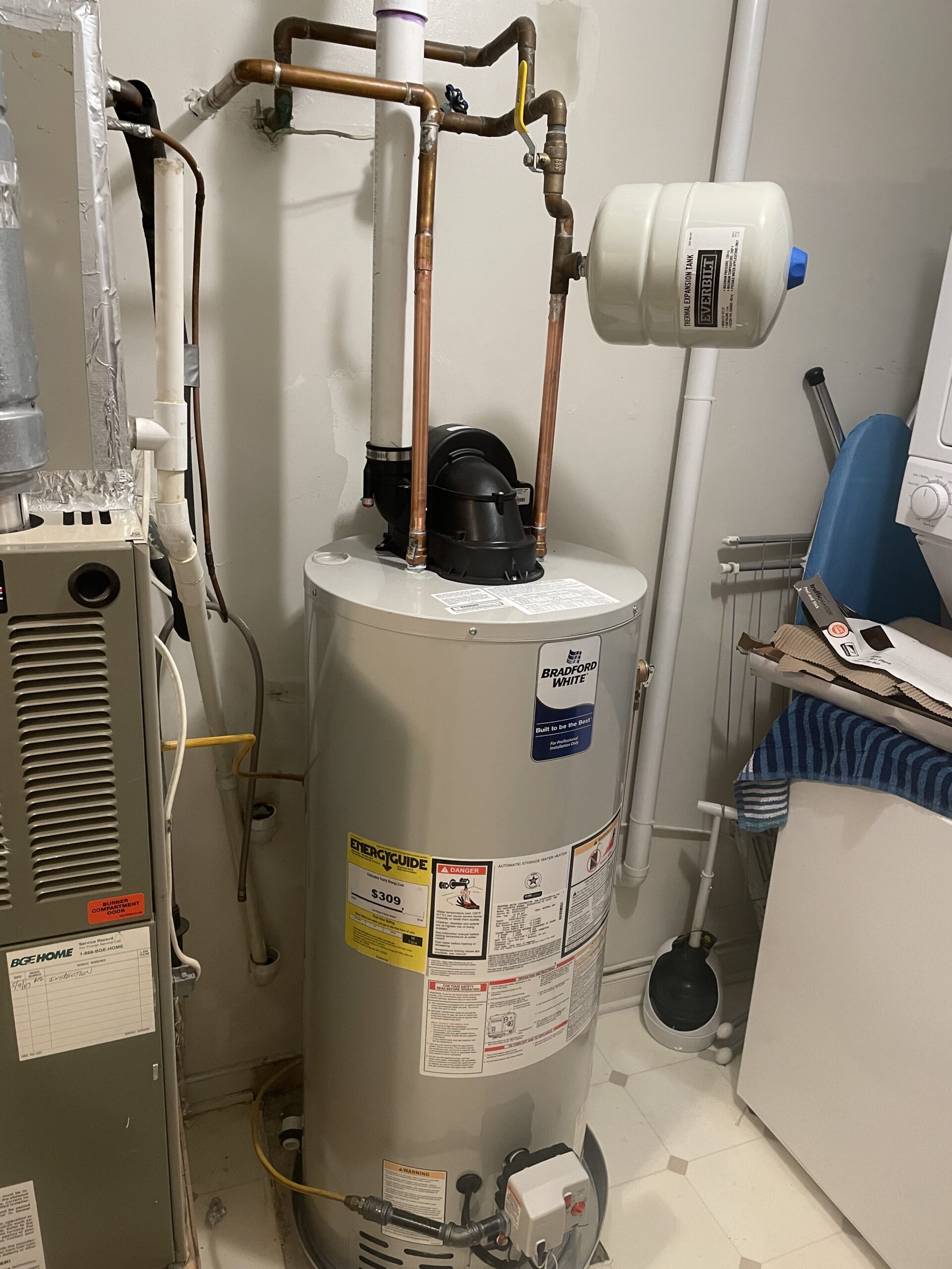 A residential water heater with attached pipes and an expansion tank is installed next to a furnace in a utility room with white walls and a tiled floor.