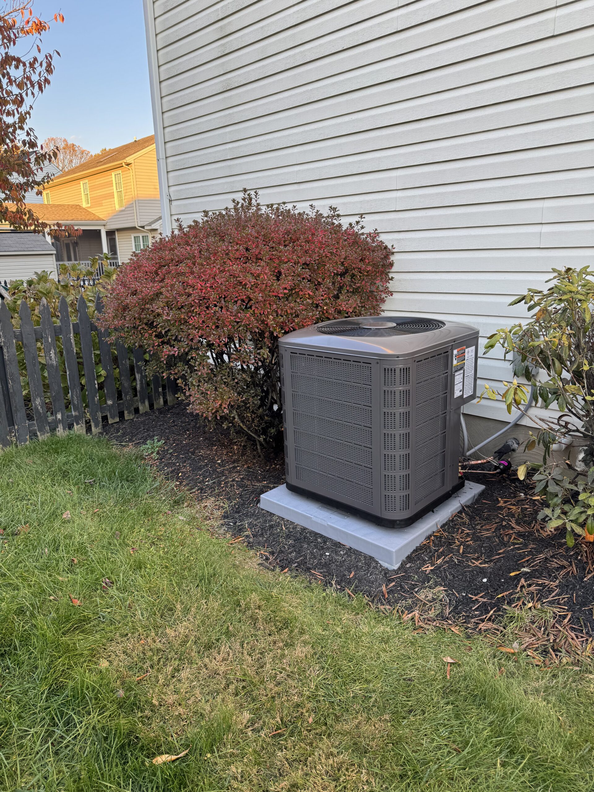 A central air conditioning unit sits on a concrete pad next to a house, surrounded by bushes and a lawn.