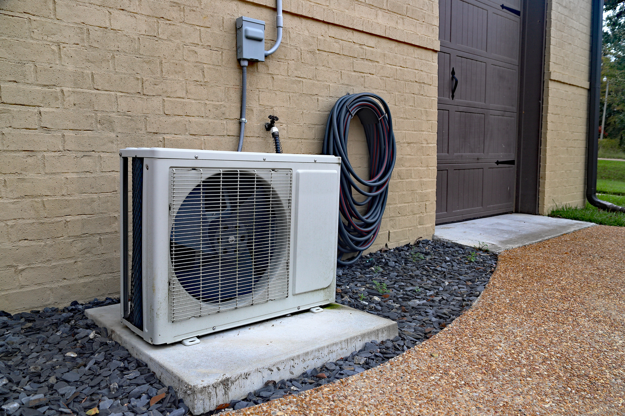 Outdoor air conditioning unit on a concrete pad next to a brick building, with coiled hoses hanging on the wall nearby.