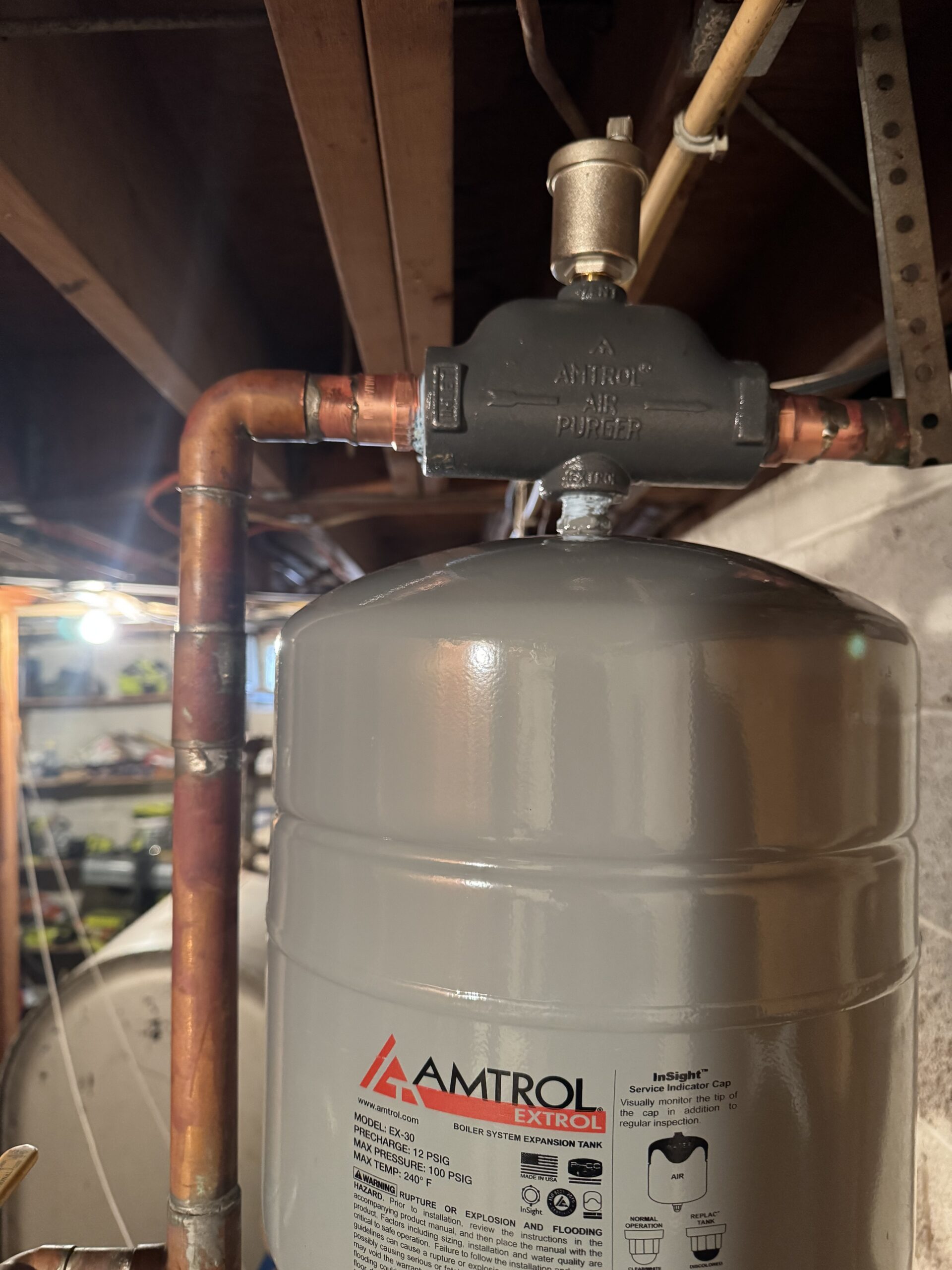 Close-up of an Amtrol expansion tank connected to copper pipes and a pressure relief valve in a basement setting.