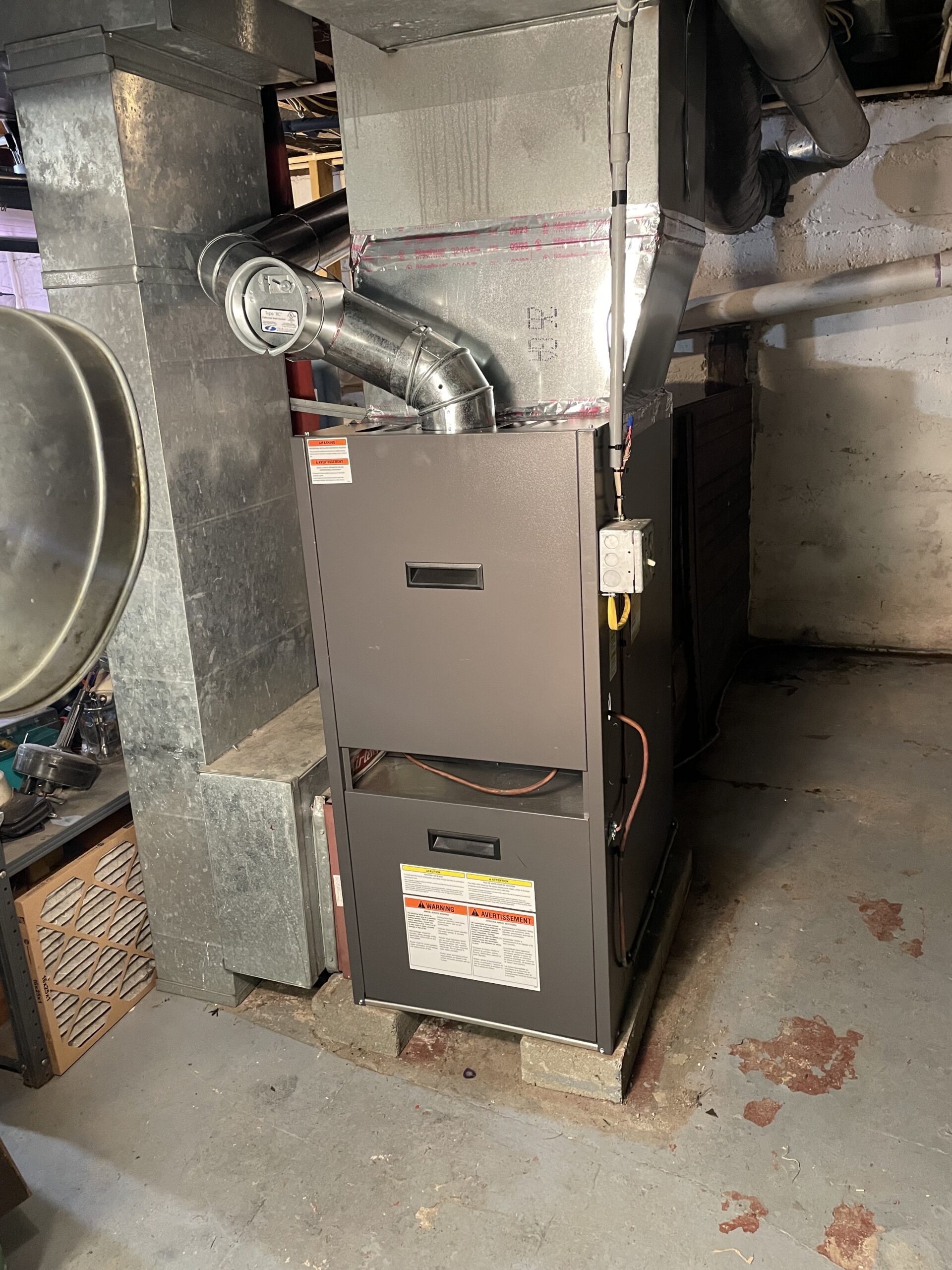 A modern forced-air furnace unit installed in a basement, connected to metal ductwork with warning labels visible on the front panels.